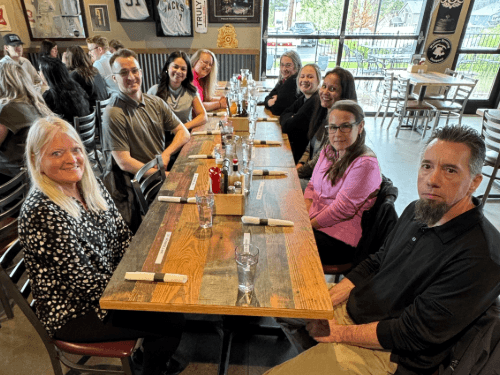 A large group of smiling dealership employees gathered around a long wooden table for a group lunch together at a restaurant.