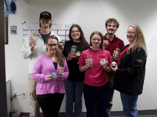 Six smiling employees standing in an office holding personalized "Thank You" glass tumblers and small crocheted animals.
