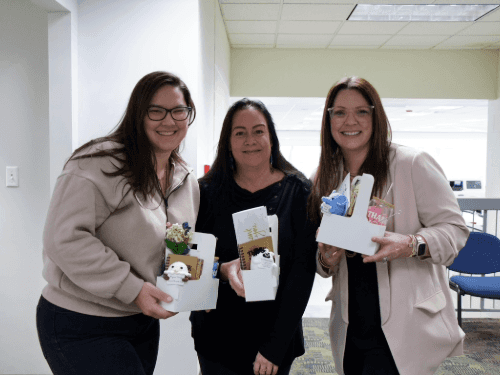 Three smiling women standing together in a hallway, holding white gift baskets filled with notebooks, tumblers, and small crocheted animals.