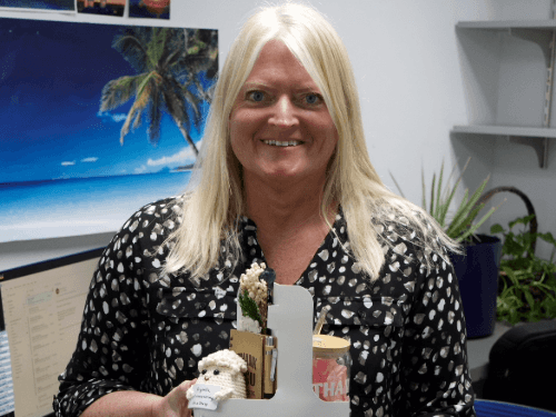 A smiling blonde woman holding a white gift basket with a small crocheted sheep, a tumbler, and a notebook in an office setting.