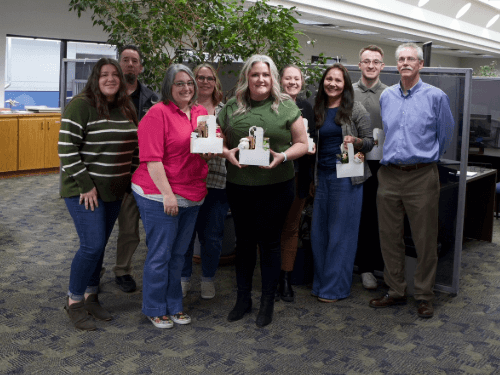 A group of smiling dealership employees standing together in an office showroom holding white administrative gift baskets.
