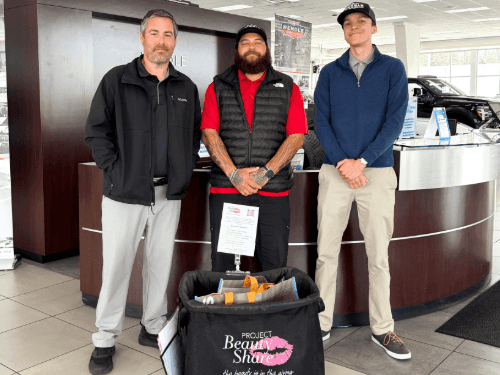 Three men standing by a showroom desk with a Project Beauty Share donation bin filled with bags at Wendle Ford car dealership.