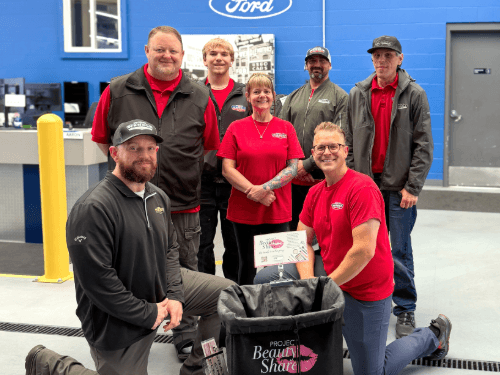 Seven smiling dealership employees posing together in a service bay around a Project Beauty Share donation bin.
