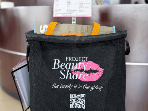 A black Project Beauty Share donation bin sitting in front of a Wendle dealership counter, holding filled shopping bags.