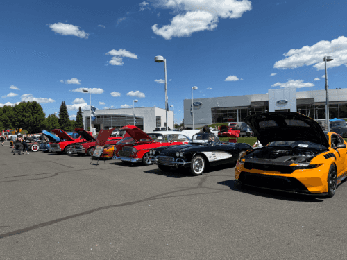 Classic cars on display at a dealership car show, including a yellow Mustang, a black Corvette, and a red coupe.