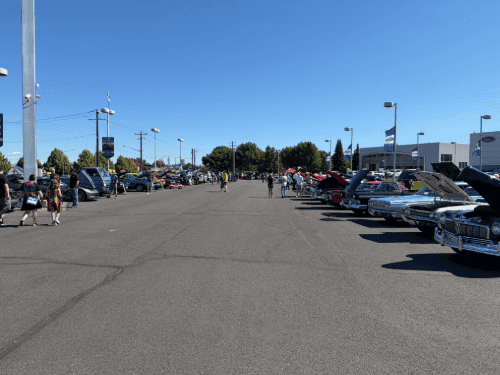 Rows of classic cars parked on an open asphalt lot at the Wendle car show, with visitors walking around under a blue sky.