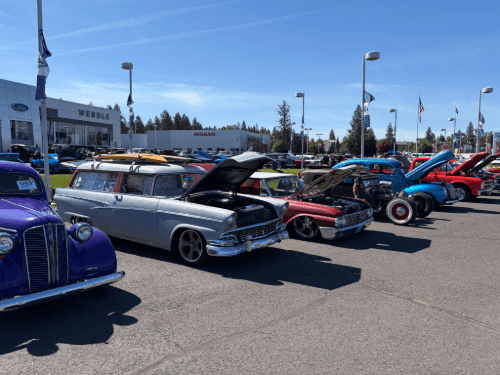 Classic cars, like a grey station wagon with surfboards on top, parked at the Wendle dealership lot under a clear blue sky.