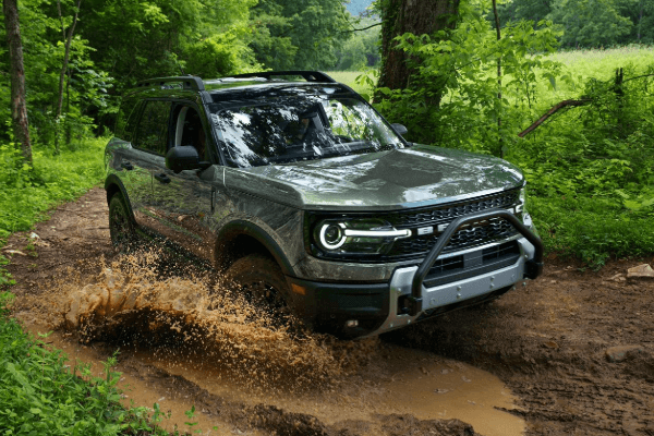 A green Ford Bronco Sport splashing muddy water as it drives through a deep puddle on a dirt trail surrounded by dense, lush green forest.