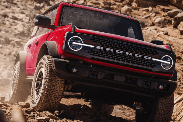 A red Ford Bronco navigating a rugged, rocky desert trail, showcasing its front grille and aggressive off-road tires.