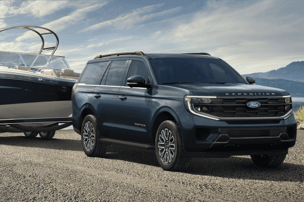 A dark blue Ford Expedition Platinum towing a large boat on a trailer along a gravel shore with water and mountains in the background.