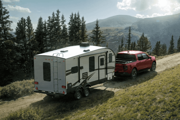 A red midsize Ford Maverick pickup truck towing a white travel trailer camper up a winding dirt mountain road under a bright, partly cloudy sky.