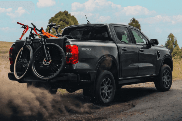 A dark grey Ford Ranger pickup truck kicking up a cloud of dust as it drives down a dirt road with two mountain bikes mounted over the tailgate pad.