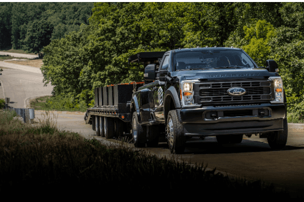 A front-angle view of a dark blue Ford Super Duty dually truck towing a heavily loaded, multi-axle flatbed trailer down a paved road with green trees in the background.