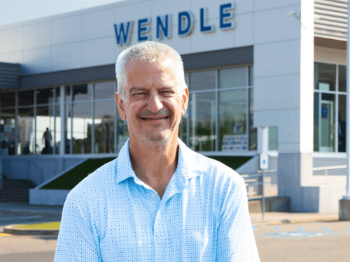 Ken smiling outside the Wendle dealership building in a light blue patterned polo shirt and jeans for his Wendleversary.