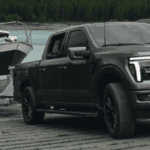 A dark grey Ford F-150 pickup truck pulling a boat on a trailer down a boat launch ramp near a lake.