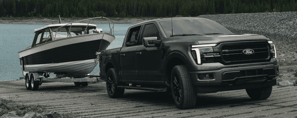 A dark grey Ford F-150 pickup truck pulling a boat on a trailer down a boat launch ramp near a lake.